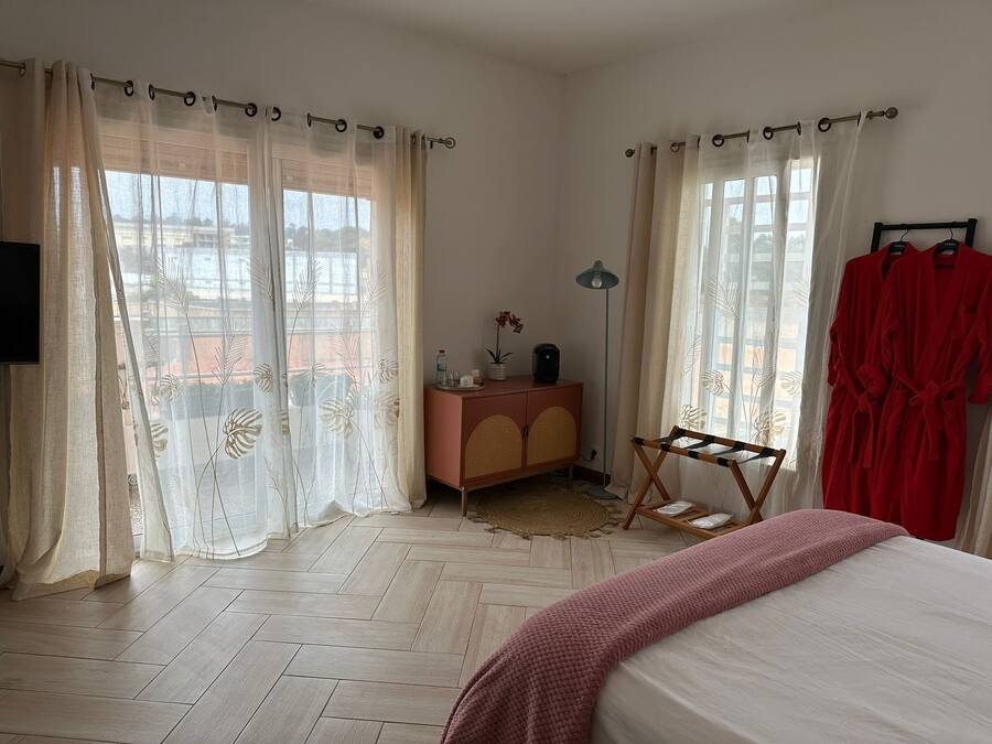 Quarto com cama, cômoda, espelho, janela com cortinas leves e roupão vermelho pendurado em um suporte.