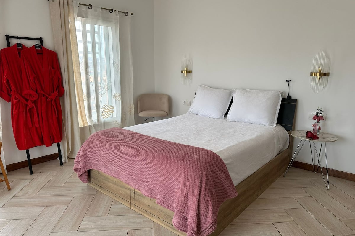 A bedroom with a bed, a chair, a small table, and a robe rack.