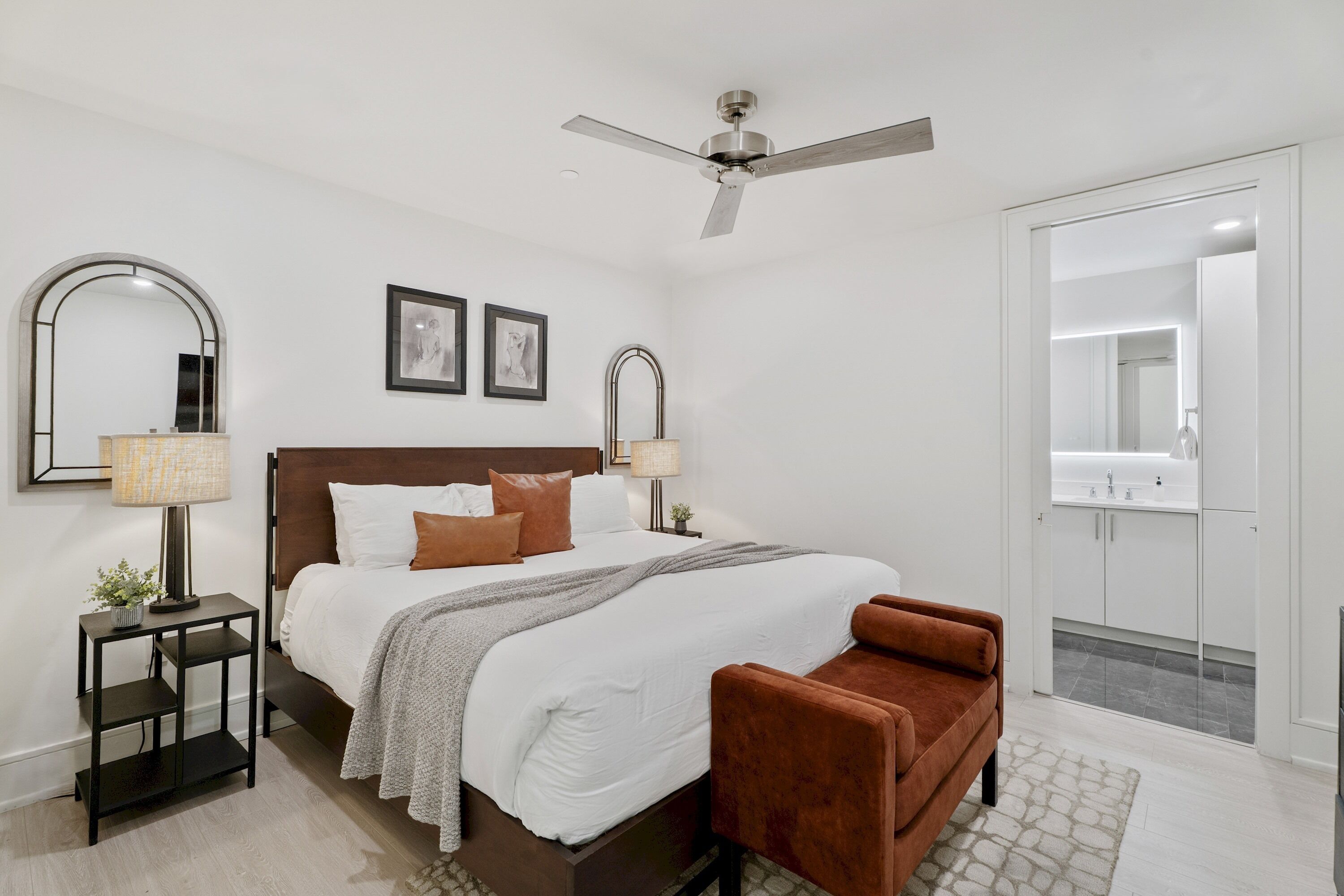 A hotel room with a bed, a ceiling fan, a nightstand, a chair, a mirror, and a bathroom visible through an open door.