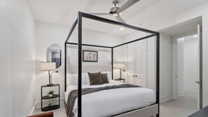 Uma cama com dossel, um ventilador de teto, luminárias de cabeceira, uma pequena planta e um quadro na parede.
