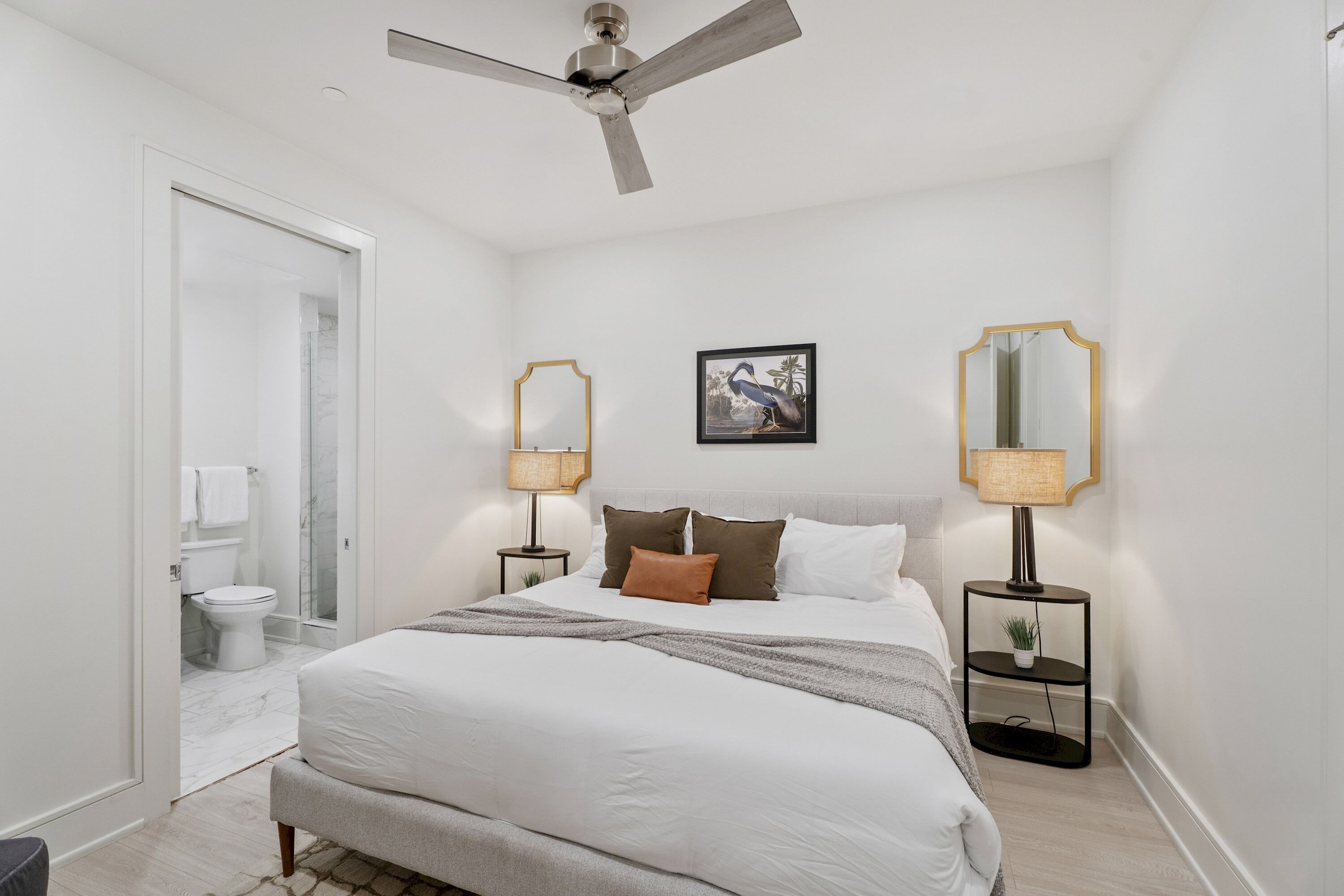 Une chambre à coucher avec un lit, deux tables de chevet avec des lampes, un ventilateur de plafond, un tableau encadré au mur, et une salle de bain avec des toilettes et une douche.