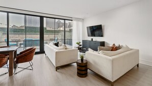Uma sala de estar moderna com uma janela ampla, uma TV de tela plana, dois sofás, uma mesa de jantar e uma pequena mesa lateral.