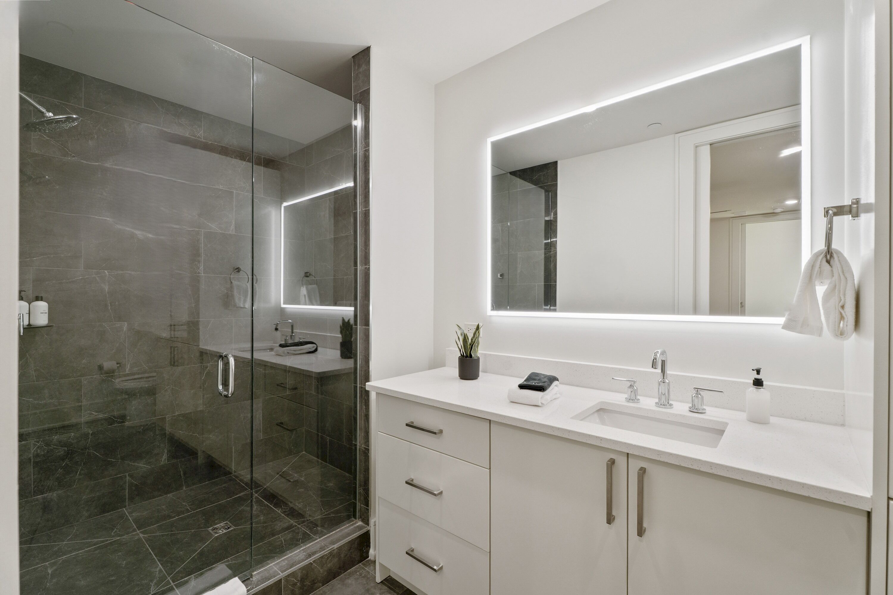 Un baño moderno con ducha envuelta en cristal, tocador blanco con un espejo grande y un lavamanos.