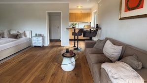 En moderne stue med sovesofa, glasbord og et køkken i baggrunden.