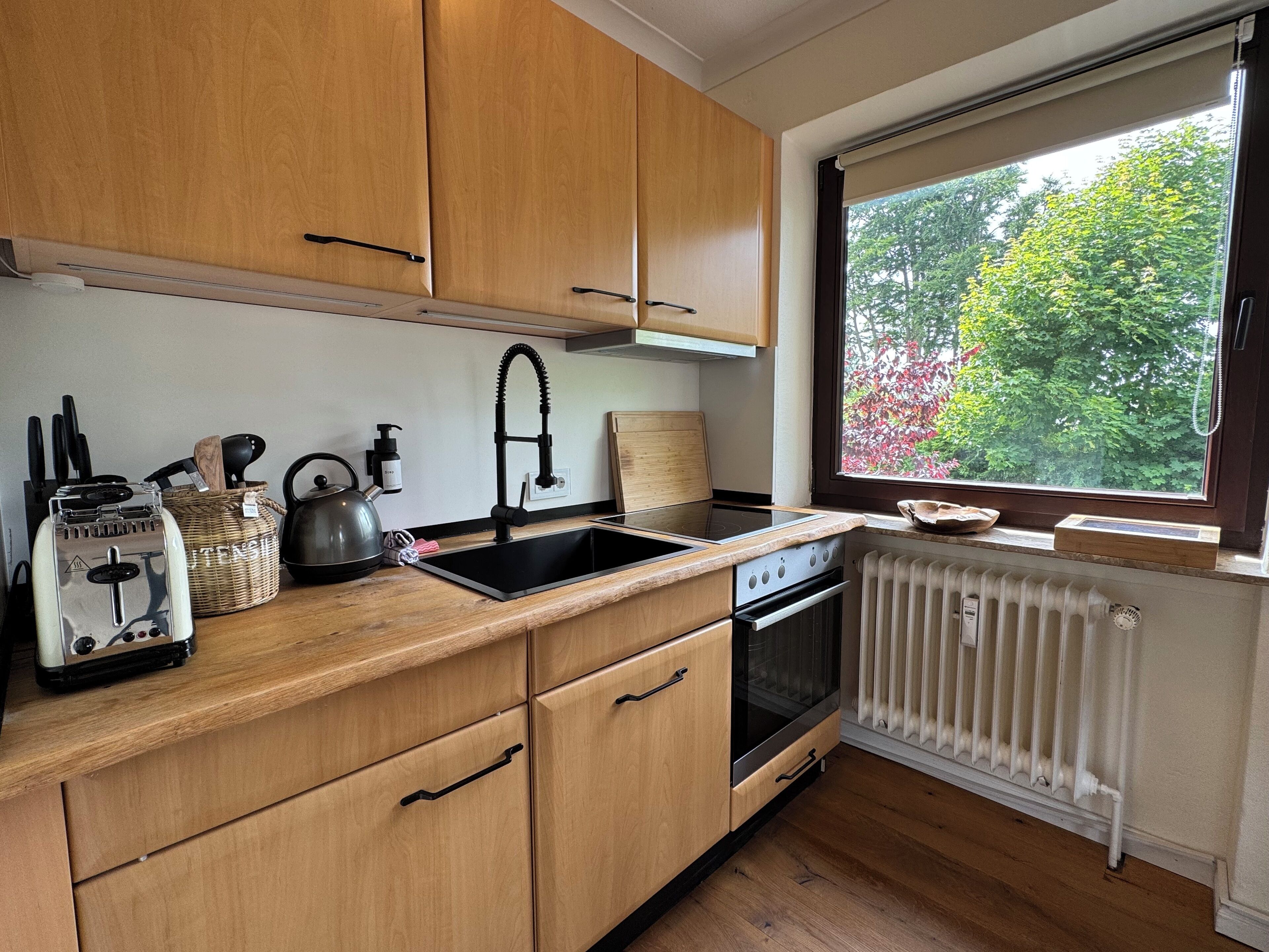 Eine Küche mit Holzausstattung, einem schwarzen Wasserhahn, einem Wasserkocher, einem Toaster und einem Fenster mit Blick auf Bäume.