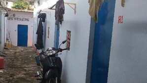 Un couloir étroit, avec une moto garée sur le côté gauche. Les murs sont peints en blanc avec des touches de bleu, et une porte rouge porte l’inscription « H1 ».