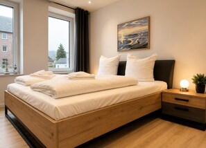 A modern bedroom with a wooden bed, white linens, a nightstand with a lamp, and a painting on the wall. - House Deichlust Whg.1,Strichweg 165, 27476 Cuxhaven-Döse (Cuxhaven-Döse)