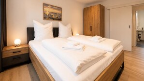 A double bed with white linens, a nightstand with a lamp, a wooden wardrobe, and a painting on the wall. - House Deichlust Whg.1,Strichweg 165, 27476 Cuxhaven-Döse (Cuxhaven-Döse)