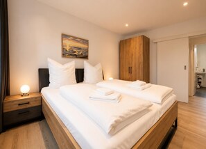 A double bed with white linens, a nightstand with a lamp, a wooden wardrobe, and a painting on the wall. - House Deichlust Whg.1,Strichweg 165, 27476 Cuxhaven-Döse (Cuxhaven-Döse)