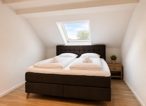 A bedroom with a dark headboard, a skylight, a bedside table with a plant, and neatly folded towels.