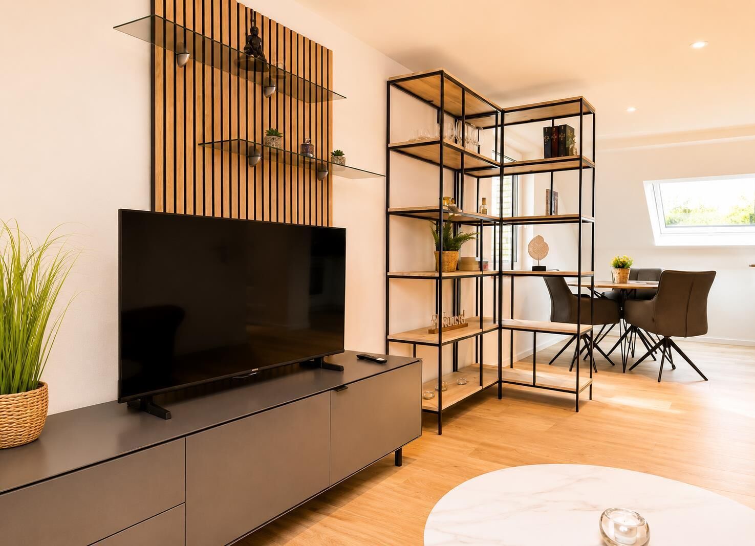 A modern living room with a flat-screen TV, a wooden slat wall, a glass shelving unit, a dining area with a table and chairs, and a potted plant.