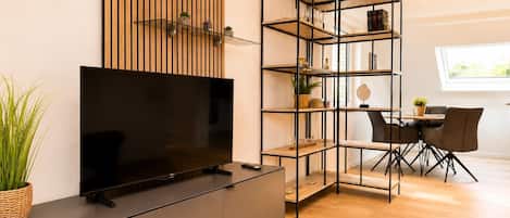 A modern living room with a flat-screen TV, a wooden slat wall, a glass shelving unit, a dining area with a table and chairs, and a potted plant.