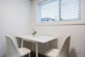 Une petite pièce blanche, avec une seule table et deux chaises, une fenêtre avec des stores et une petite plante en pot posée sur le rebord de la fenêtre.