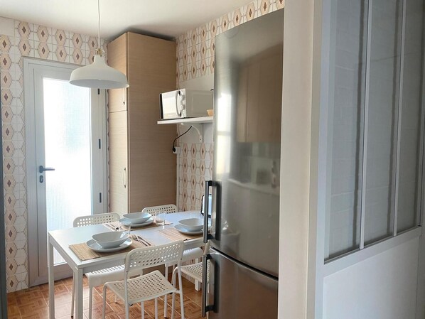 Cocina compacta con refrigerador de acero inoxidable, una mesa de comedor con sillas blancas y una lámpara colgante.
