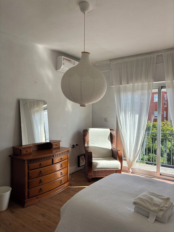 A bedroom with a wooden dresser, a wicker chair, a large bed, a hanging light fixture, and a view of a red building through sheer curtains.