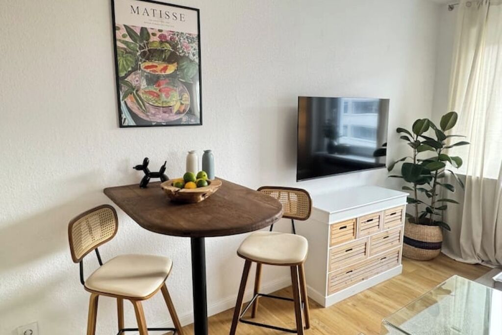 Una sala moderna con una mesa redonda para comer, dos sillas, un televisor de pantalla plana, una planta grande en maceta y una obra de arte colgada en la pared.