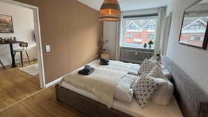 Un dormitorio con cama, lámpara colgante, ventana con vista a edificios y una pequeña mesa con sillas.
