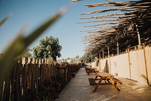 A pathway lined with wooden benches and tables, leading to a covered area with a thatched roof. - DUNIA Desert Suites (Mitzpe Ramon)