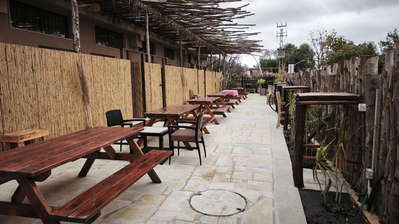 An outdoor dining area with wooden tables and chairs, a bamboo wall, and a stone floor.