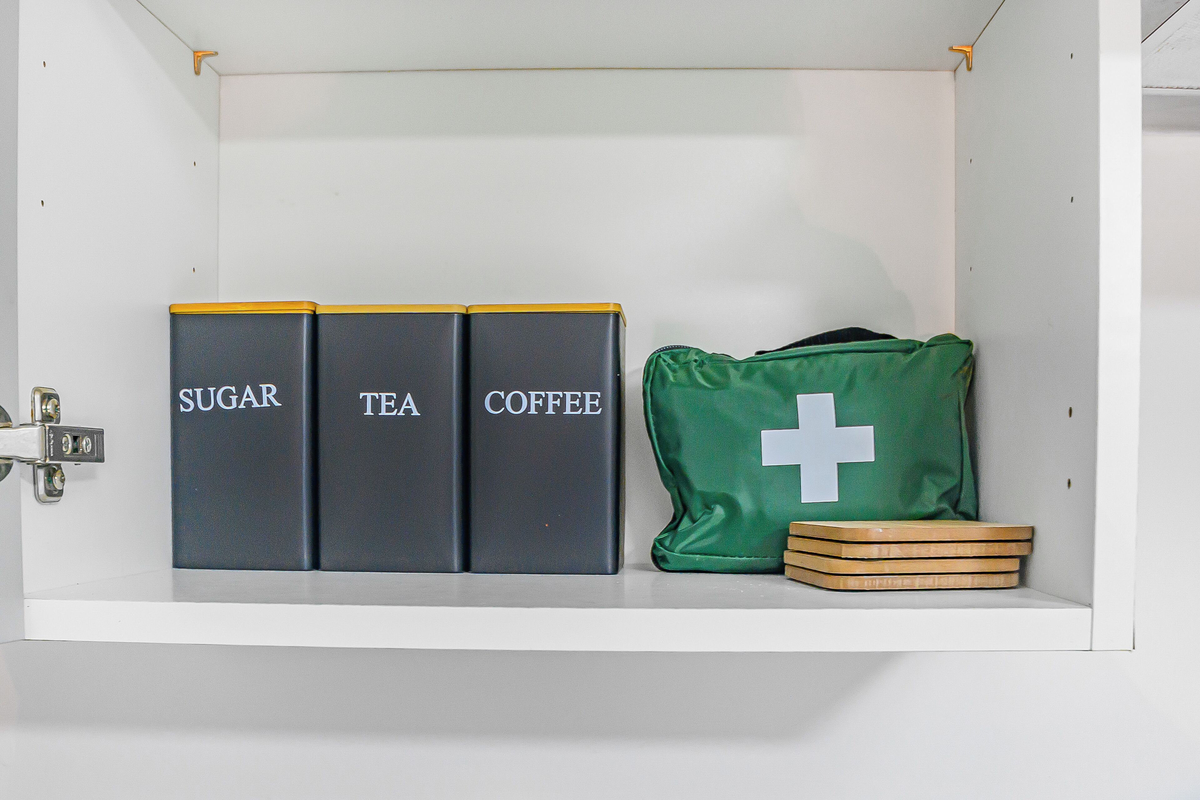 Un área de almacenamiento con contenedores etiquetados para azúcar, té y café, y una bolsa verde con una cruz blanca.