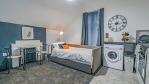 A compact bedroom with a bed, a washing machine, a small table with chairs, and a clock on the wall. - Modern 1 Bed - Near City Centre  (Manchester)