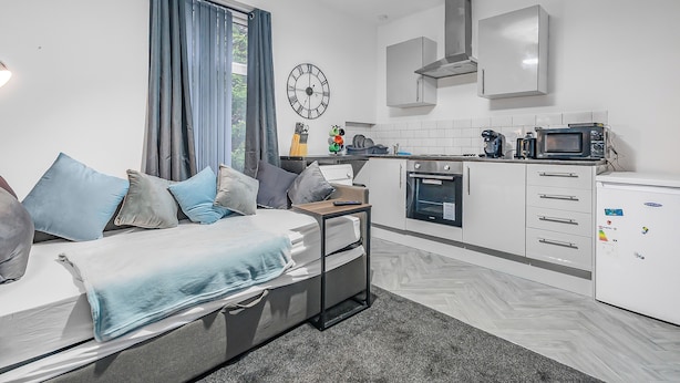 A compact living space with a bed, a small table, a kitchen area including a microwave, and a clock on the wall. - Modern 1 Bed - Near City Centre  (Manchester)