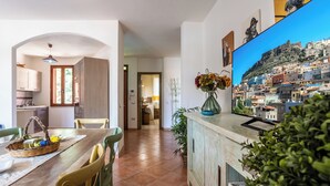 Un coin repas avec une table en bois, des chaises et un panier de fruits. Un téléviseur fixé au mur diffuse des images d’un paysage urbain.