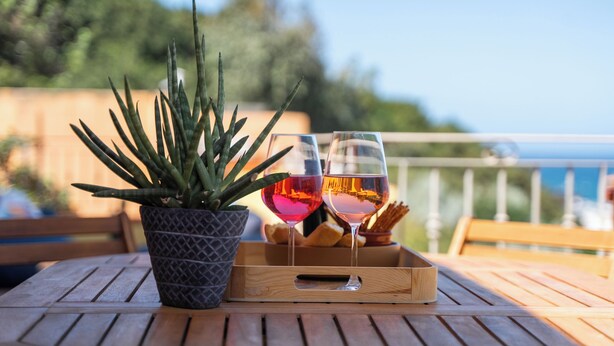 Une table en bois, ornée d’un pot de fleurs et de deux verres de vin rouge, est placée sur un balcon d’où l’on peut admirer la mer.