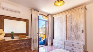 Une chambre à coucher avec une commode en bois, un grand miroir, un lit recouvert d’une couvre-lit à motifs et une armoire en bois clair.
