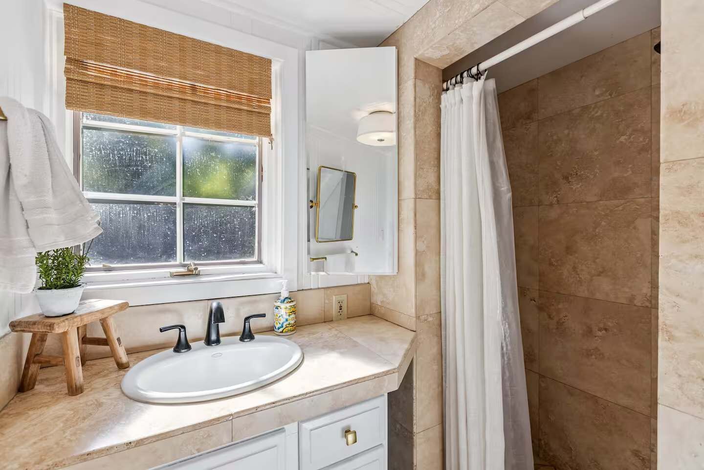 A bathroom with a large window, a sink with three faucets, a mirror, a towel rack, and a shower curtain.