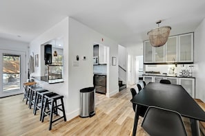 A modern kitchen with a dining area, featuring a long table, bar stools, and a hanging light fixture. - Spacious 7-Bedroom Compound w 2 Large POOLS, Fire-pit, Grill & fully-fenced yard (Austin)