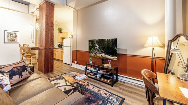 Un salon comprenant un canapé, une table basse et un téléviseur. Il y a également un coin repas avec une table et des chaises. La pièce est dotée d’un parquet et d’un grand tableau accroché au mur.