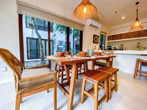 A dining area with a wooden table and chairs, a woven ceiling light, and large windows with a view of trees and buildings outside. - Selva Norte Nature Retreat with jacuzzi (Tulum)