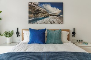 A bedroom with a bed, pillows, a bedside table with a plant, a wall-mounted lamp, and a framed picture of a coastal landscape. - Orange Suite at Jellybean House (St. John's)