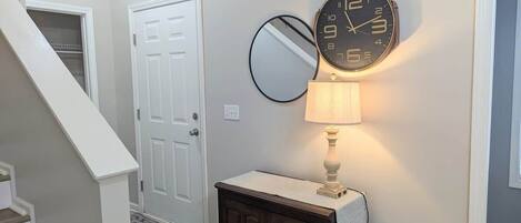 Un pasillo con una escalera, una puerta blanca, un armario de madera oscura con un paño a rayas, un espejo redondo montado en la pared y un reloj de pared.