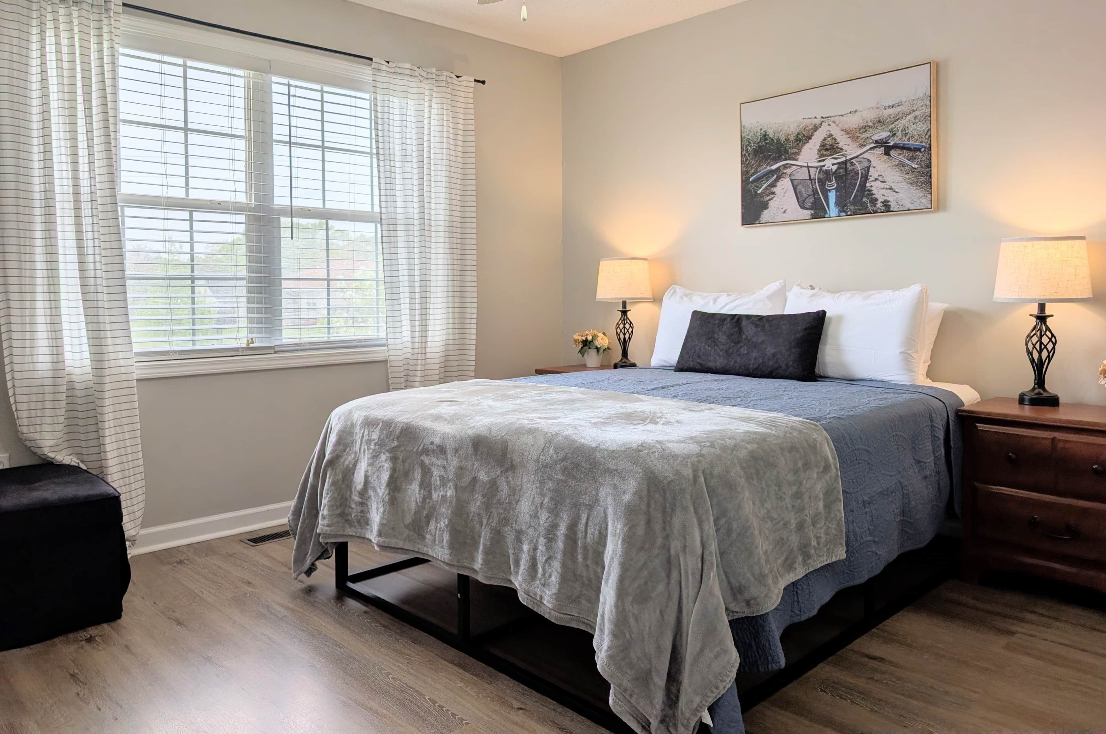 Un dormitorio con cama, dos lámparas de noche, una mesita de noche, una ventana con cortinas y un cuadro en la pared.