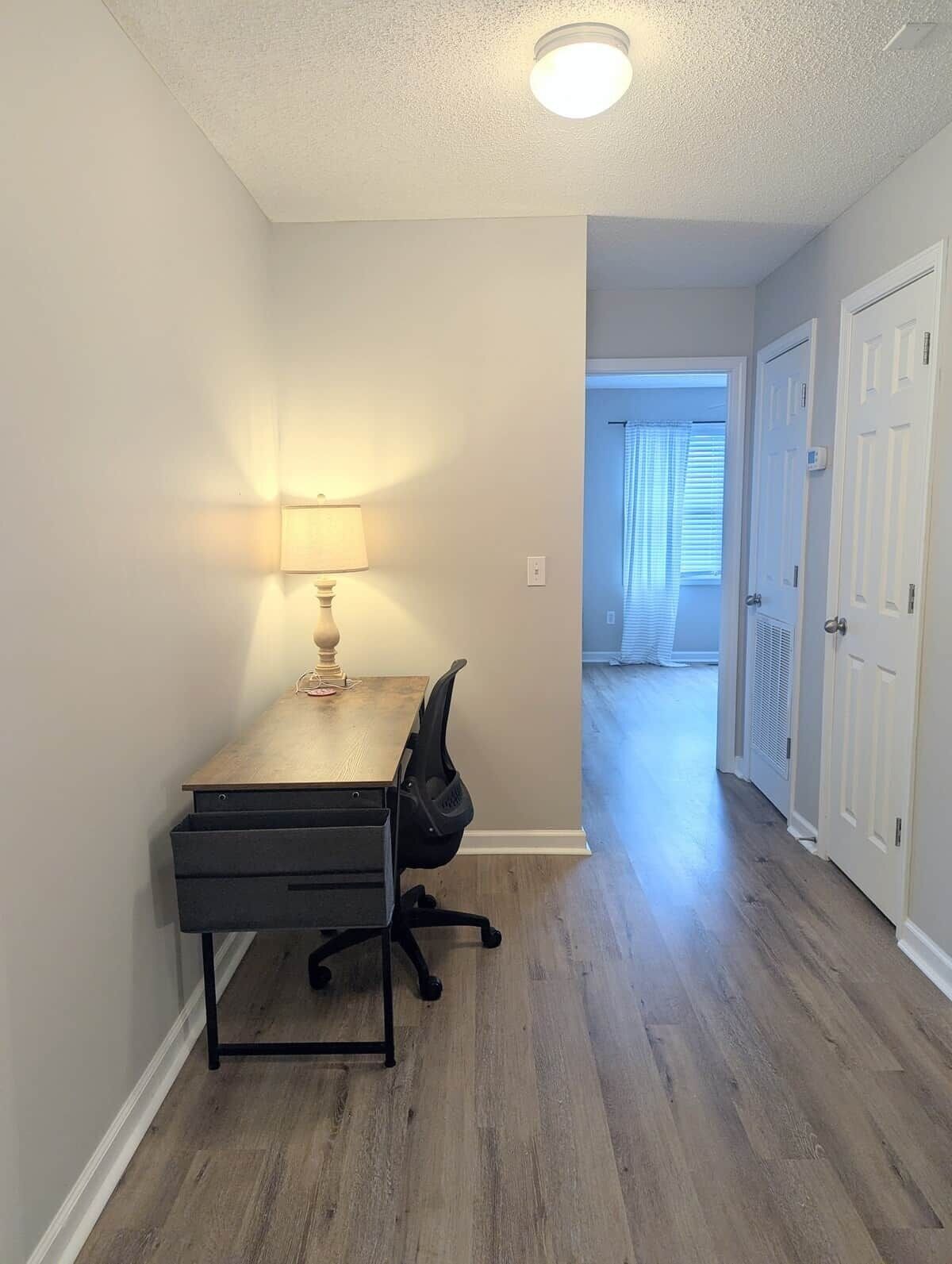 Una habitación con escritorio, silla, lámpara y un pasillo que conduce a otra habitación.