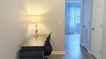 Una habitación con escritorio, silla, lámpara y un pasillo que conduce a otra habitación.