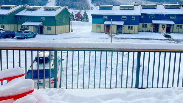 A snow-covered building complex with multiple units, a parking area, and a view of a forested hill in the background. - Alpine Retreat - Ski & Golf Across From Hill - Full Townhouse - Fully Stocked (Ellicottville)