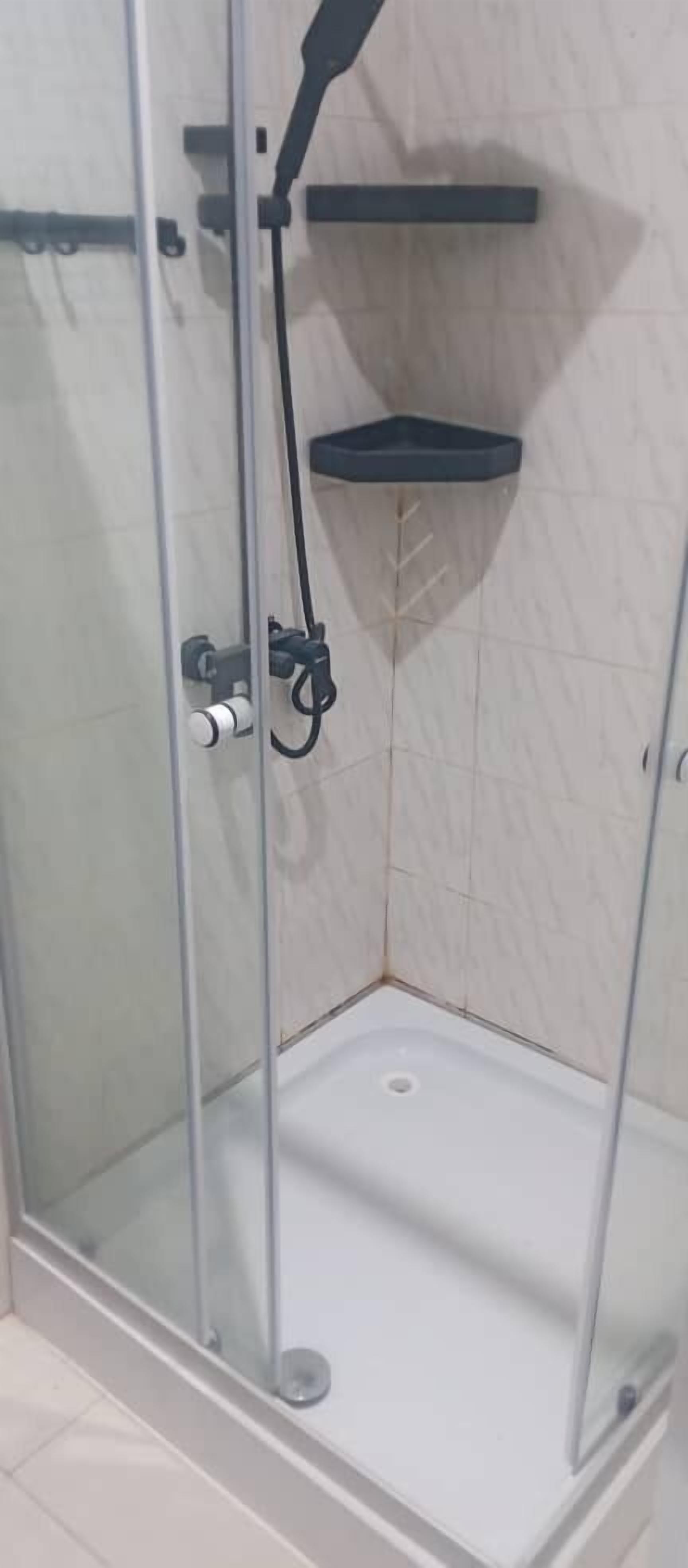 A glass-enclosed shower stall with a white floor, a black showerhead, and a black shelf on the wall.