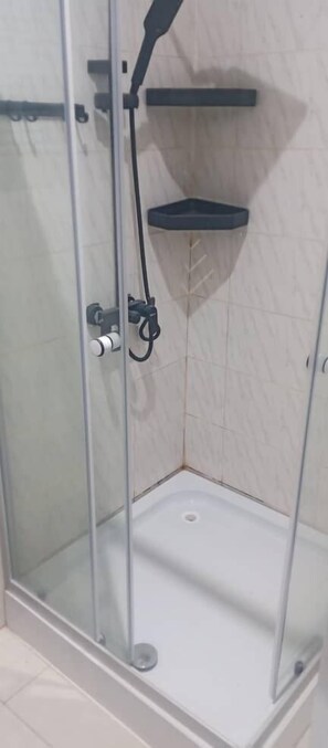 A glass-enclosed shower stall with a white floor, a black showerhead, and a black shelf on the wall. - SNS Apartments (Adentan)