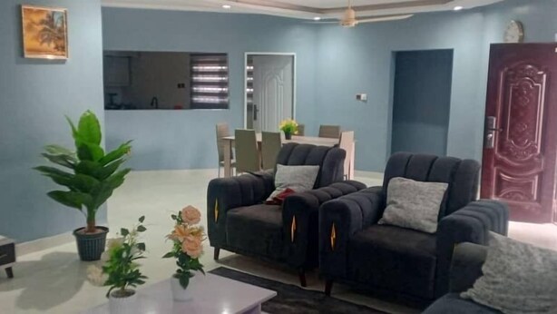 A living room with a sofa set, a coffee table, a TV, and a dining area in the background. - SNS Apartments (Adentan)