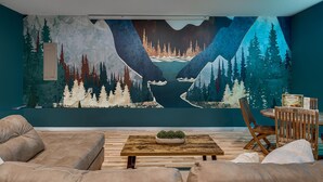 Una sala de estar con un gran mural de un bosque y montañas, una mesa de centro de madera y un comedor con una silla y mesa de madera.