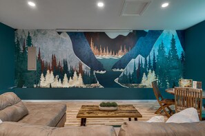 A living room with a large mural of a forest and mountains, a wooden coffee table, and a dining area with a wooden chair and table. - Peaceful Riverfront Gem + SNP Mountain Magic w/ Hot Tub & 6-8 Person Sauna (Shenandoah)