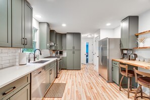 A modern kitchen with grey cabinets, a stainless steel refrigerator, and a wooden dining table. - Peaceful Riverfront Gem + SNP Mountain Magic w/ Hot Tub & 6-8 Person Sauna (Shenandoah)