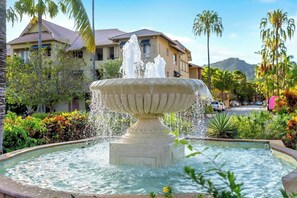 Una fuente con chorros de agua, rodeada de vegetación y un edificio al fondo.