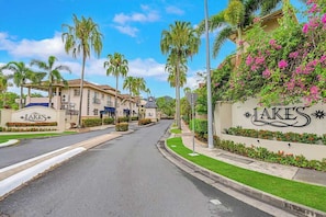 Une vue d’une rue d’un quartier résidentiel, avec un panneau indiquant « LAKES » et des palmiers bordant la rue.