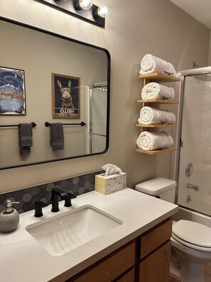 A bathroom with a large mirror, a sink with a soap dispenser, a toilet, and a glass-enclosed shower. There are towels on a shelf and a tissue box on the counter. - The New Minglewood - a Beautiful 4-bedroom house with Hot Tub (Norden)