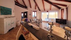 Een ruime woonkamer met houten plafond, een eettafel met stoelen, een bank, een televisie en een groot raam met gordijnen.