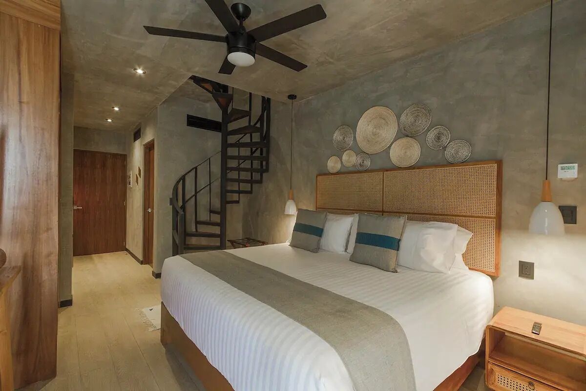 A bedroom with a large bed, a ceiling fan, a rattan headboard, and a staircase in the background.